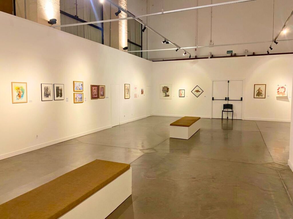 Grande galeria de arte com paredes brancas e iluminação direcionada. O espaço está lotado de visitantes que observam obras expostas nas paredes e em painéis centrais. O ambiente é vibrante e acolhedor.