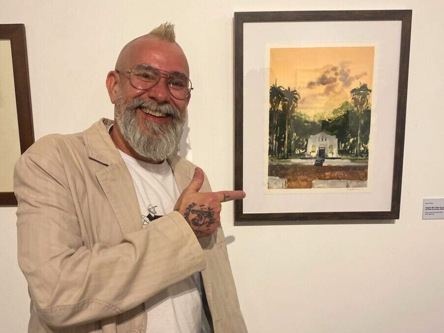 Homem branco, careca, de terno azul escuro, toca flauta transversal em pé, em espaço interno da galeria. Ao fundo, músicos de apoio e caixas de som. À direita, há um saxofone encostado na parede.