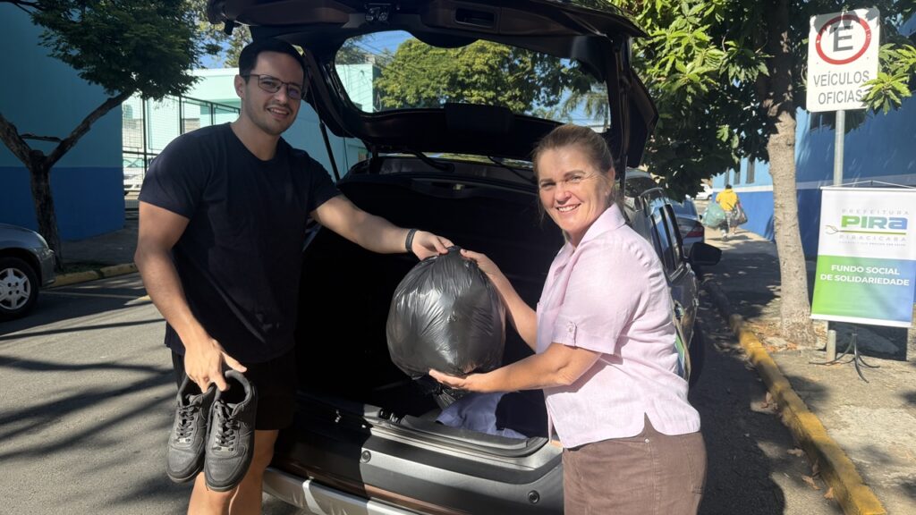 A imagem mostra dois indivíduos, um homem e uma mulher, perto de um carro com a mala aberta. O homem está vestindo uma camiseta preta e calções escuros. Ele está segurando um par de tênis. A mulher está vestindo uma blusa rosa e calças marrons. Ela está segurando um saco de lixo preto. O carro é uma SUV escura. Ao fundo, há um prédio azul e uma placa com a inscrição "Veículos Oficiais". Há também uma placa com o logotipo da Prefeitura de Piracicaba e a inscrição "Fundo Social de Solidariedade". O cenário parece ser uma rua ou estacionamento. A imagem sugere uma doação de itens para uma organização de caridade.  A iluminação indica que a foto foi tirada durante o dia. 