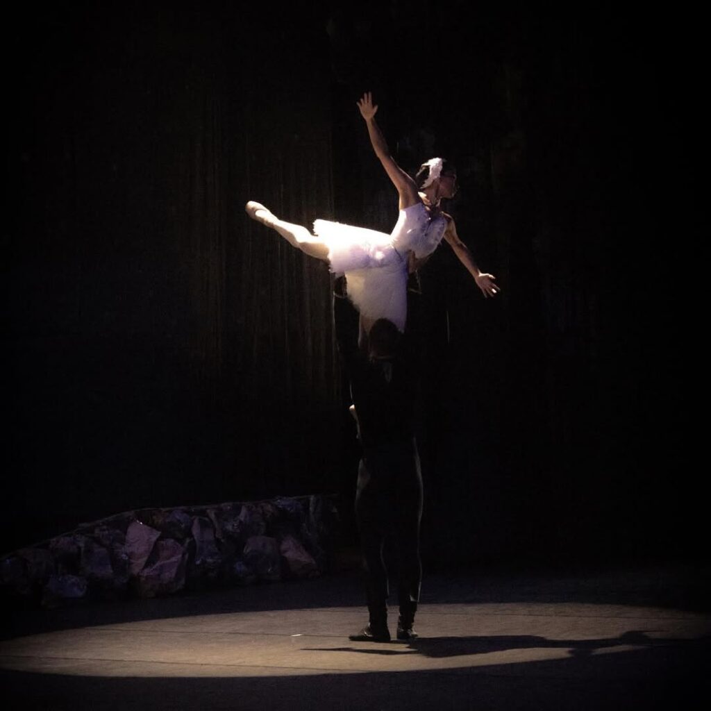 Em um palco escuro com iluminação pontual, uma bailarina vestida com figurino branco é erguida por um bailarino de roupas pretas. Ela está com os braços abertos e uma das pernas estendida para trás, em posição de voo.