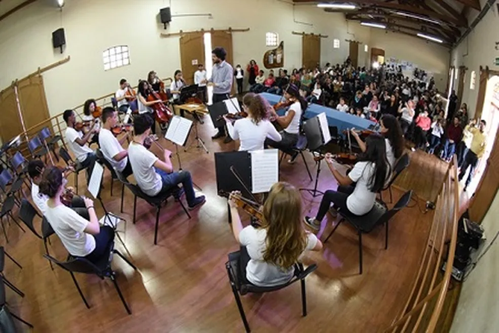 Jovens músicos tocam violinos e violoncelos em apresentação de orquestra no auditório com público ao fundo. Estão posicionados em semicírculo, usando camisetas brancas e calças escuras.