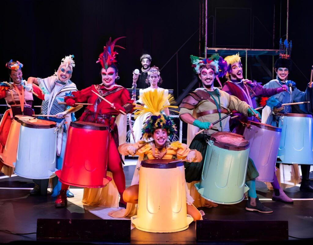 Grupo de atores do espetáculo Parará – Um Reino pela Música se apresenta no palco com fantasias coloridas inspiradas em aves e personagens lúdicos. Eles tocam tambores feitos com baldes plásticos grandes de cores vibrantes como vermelho, amarelo, verde, azul e lilás. Ao centro, uma atriz agachada sorri com expressão animada, usando fantasia amarela com penas. Todos estão maquiados artisticamente e demonstram alegria e energia em cena. O fundo é escuro, destacando as cores intensas dos figurinos.