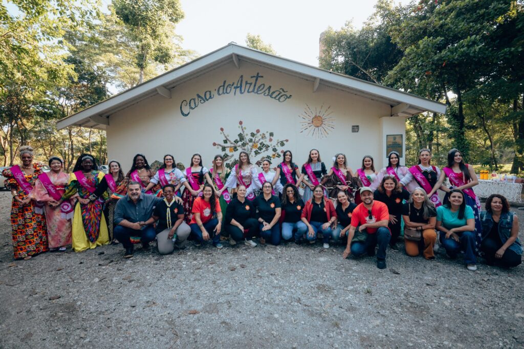 A imagem mostra um grupo de pessoas reunidas em frente a um edifício chamado "Casa do Artesão". A fachada tem um mural vibrante com uma árvore de folhas coloridas e um sol. Os indivíduos na foto vestem roupas diversas e algumas pessoas usam faixas com títulos como "Miss". O ambiente parece ser ao ar livre, cercado por árvores e vegetação.