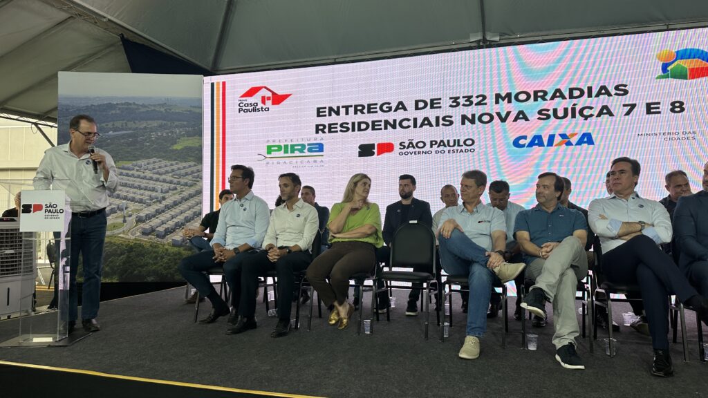 Durante a cerimônia de entrega das moradias do Residencial Nova Suíça 7 e 8, o prefeito discursou destacando a parceria com o Governo do Estado. No palco, autoridades municipais, estaduais e representantes da Caixa acompanharam o evento. O painel ao fundo reforça os programas envolvidos. O clima foi de celebração e comprometimento com a habitação.