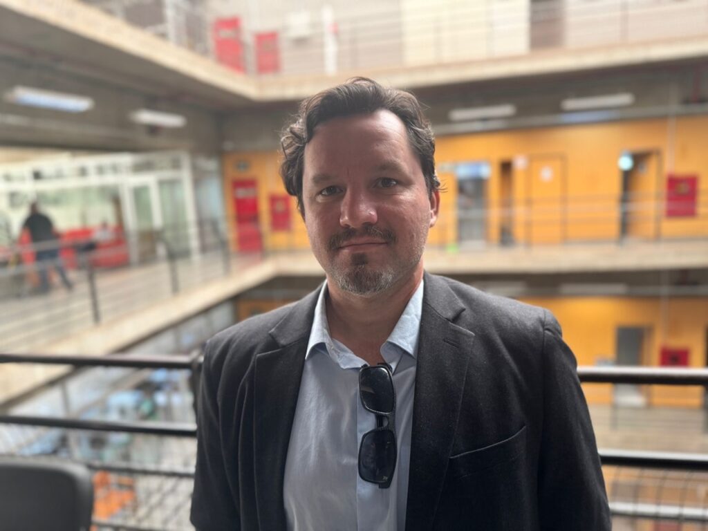 A imagem mostra um homem de meia-idade, com cabelo castanho escuro e barba aparada, vestindo um blazer escuro sobre uma camisa azul-clara. Ele está usando óculos de sol pendurados no bolso do blazer. O homem parece estar em um pátio interno de um prédio, com corredores e portas visíveis ao fundo. O fundo está levemente desfocado, focando a atenção no homem em primeiro plano. A iluminação parece natural, provavelmente luz do dia, e a atmosfera geral da foto é calma e profissional. O homem está olhando diretamente para a câmera, com uma expressão neutra e séria. O detalhe da imagem é suficiente para observar os traços faciais do homem e a textura de suas roupas. A imagem tem uma qualidade fotográfica, sugerindo que foi tirada com uma câmera de alta resolução.