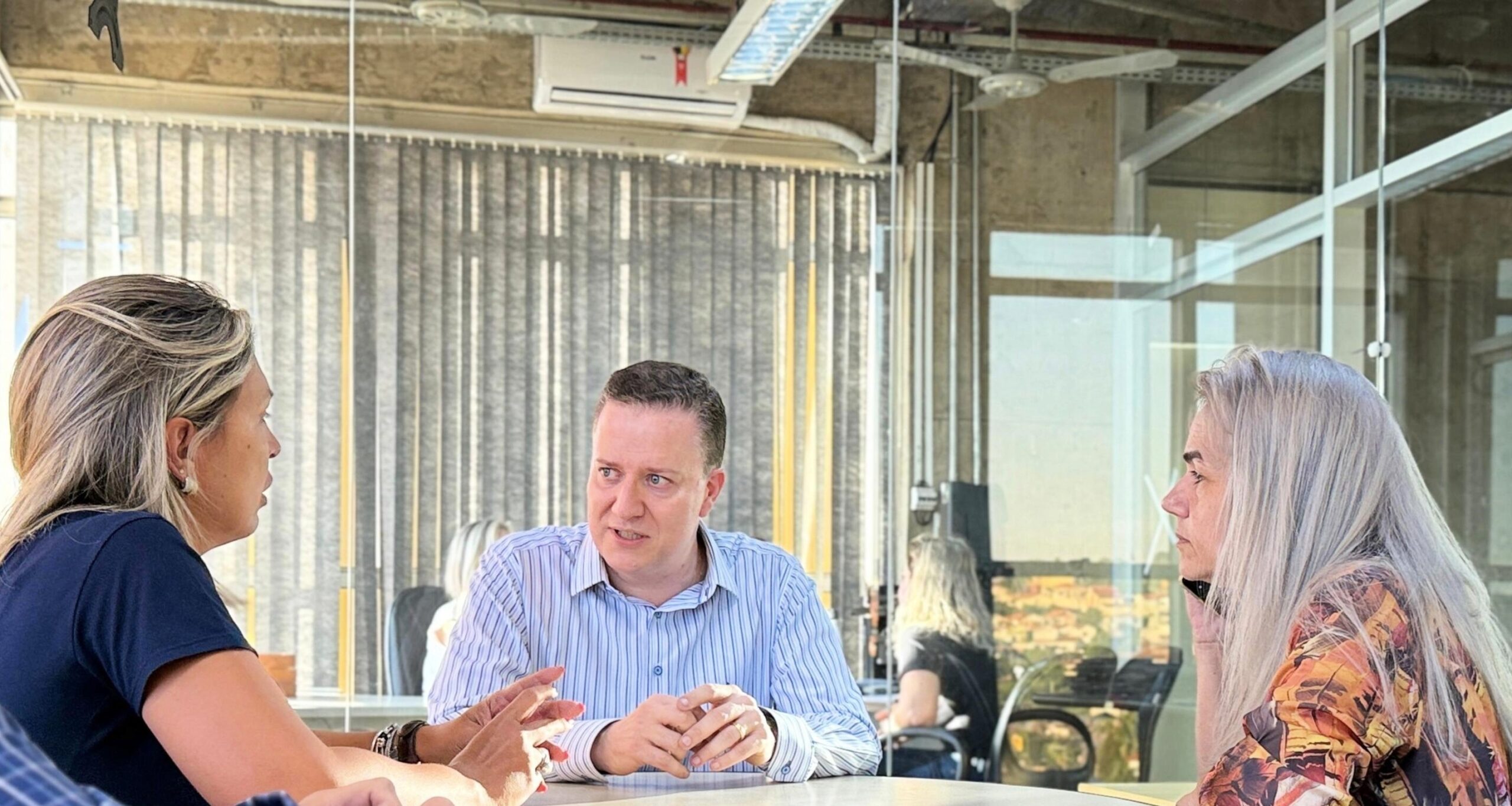 A imagem mostra quatro pessoas sentadas em torno de uma mesa de conferências, aparentemente em uma reunião de negócios. Vamos analisar os detalhes: * *Ambiente:* O local parece ser um escritório moderno, com uma grande janela de vidro que oferece uma vista externa. O espaço é bem iluminado e possui divisórias de vidro, sugerindo um ambiente de trabalho colaborativo e aberto. * *Participantes:* Há quatro indivíduos presentes: * *Homem à esquerda (de camisa xadrez azul):* Ele parece ser um dos principais participantes da reunião, possivelmente um líder ou tomador de decisão, dado sua posição e a forma como ele está envolvido na conversa. * *Mulher com cabelo loiro (à direita do homem de camisa xadrez):* Ela parece estar ativamente participando da discussão, interagindo com o homem de camisa azul e o homem sentado em frente. Sua postura sugere engajamento e atenção ao que está sendo discutido. * *Homem de camisa listrada (ao centro):* Ele está sentado de frente para as outras duas pessoas, e sua expressão e postura sugerem que ele está apresentando algo ou respondendo a perguntas. Ele parece ser um ponto focal da conversa. * *Mulher com cabelo grisalho longo (à direita, de costas para a câmera):* Ela parece estar menos envolvida na discussão principal, possivelmente ouvindo ou aguardando sua vez de falar. Ela está ao telefone ou com um dispositivo semelhante. * *Dinâmica da reunião:* A imagem sugere uma dinâmica de discussão ou brainstorming. A proximidade dos participantes, suas expressões faciais e a posição de suas mãos indicam que estão envolvidos em uma troca de ideias ativa. O homem de camisa listrada parece ser o centro da discussão, possivelmente respondendo a perguntas ou apresentando informações. * *Elementos adicionais:* Há um caderno ou bloco de notas na mesa, sugerindo que anotações estão sendo feitas durante a reunião. Em resumo, a imagem captura um momento de uma reunião de negócios em um ambiente de trabalho moderno. A dinâmica da reunião parece ser colaborativa e focada em uma discussão específica, com o homem de camisa listrada como um ponto central de informação. A mulher de cabelo grisalho parece estar em uma ligação separada, sugerindo uma possível multitarefa ou uma conversa paralela.