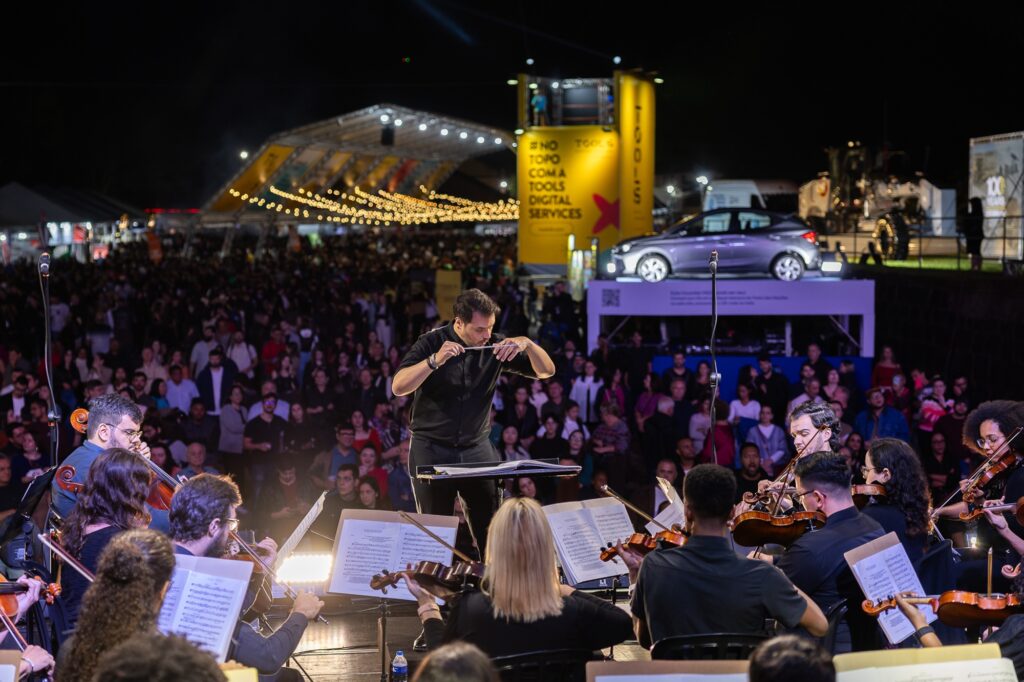 A imagem mostra uma orquestra se apresentando em um evento ao ar livre durante a noite. O maestro está conduzindo os músicos, que tocam instrumentos de cordas como violinos e violoncelos. A plateia é grande e parece estar apreciando a apresentação. Ao fundo, há uma estrutura iluminada com luzes decorativas e um grande banner amarelo.