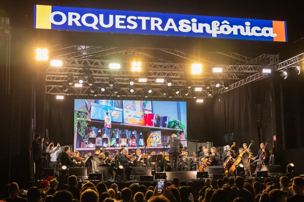 A imagem mostra uma apresentação ao vivo de uma orquestra em um palco iluminado por diversas luzes brilhantes. No fundo do palco, há um grande telão exibindo imagens coloridas, incluindo garrafas e recipientes. Acima do palco, há uma placa com o texto "ORQUESTRA Sinfônica" em letras destacadas. O público está visível em primeiro plano, assistindo à apresentação.