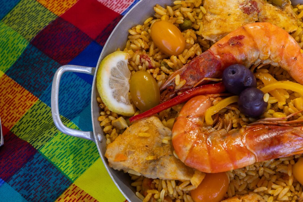 A imagem mostra uma colorida paella de frutos do mar, servida em uma panela metálica sobre uma toalha xadrez vibrante. O prato contém arroz temperado, camarões, peixe, pimentões, azeitonas roxas, tomates amarelos e uma fatia de limão. É uma especialidade típica da culinária espanhola. A apresentação é rica e apetitosa.