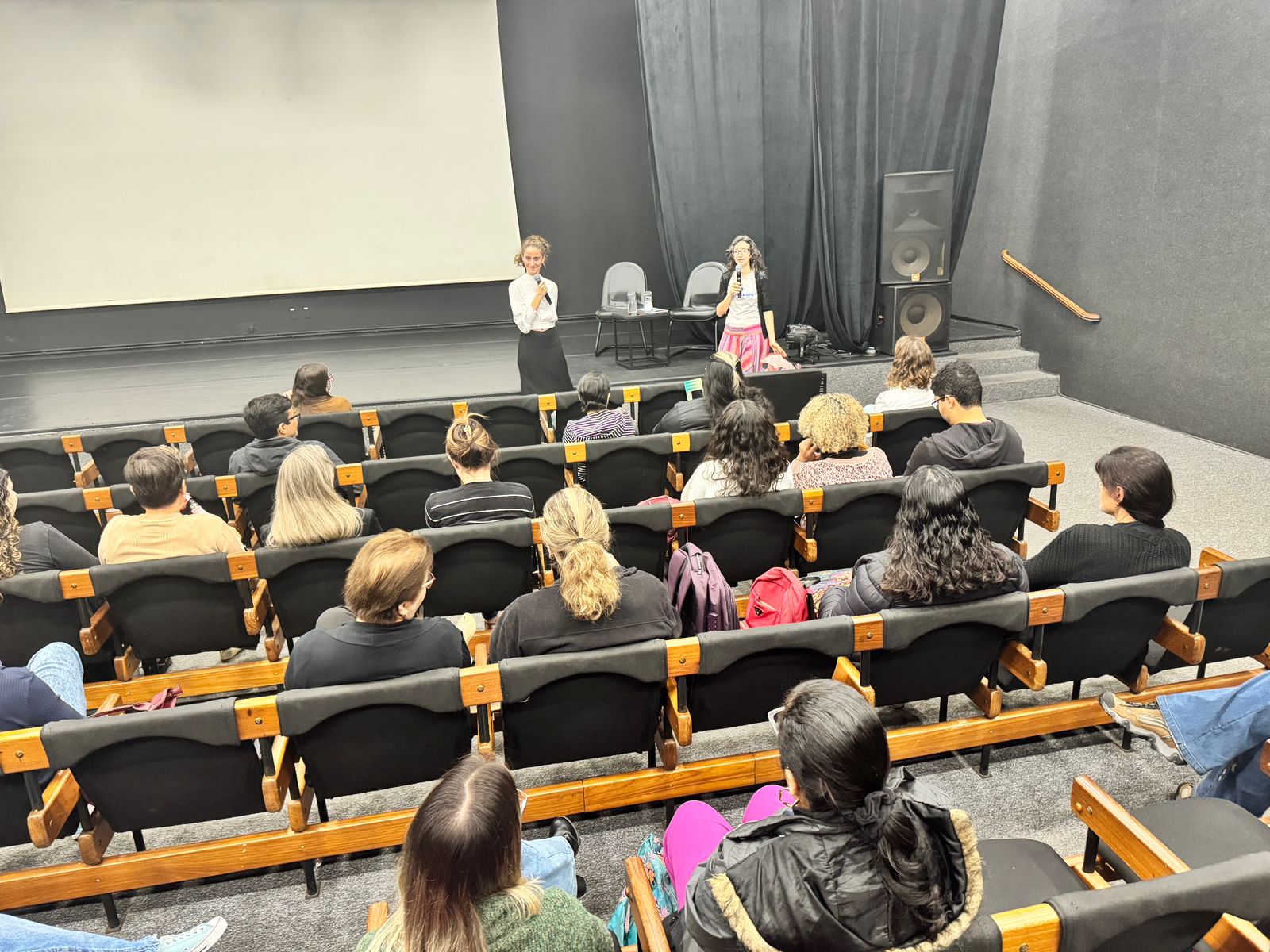 A imagem mostra uma apresentação numa sala de cinema ou auditório. Duas mulheres estão no palco, uma com um microfone, aparentemente a dar uma palestra ou apresentação para uma audiência sentada em cadeiras individuais. A plateia é composta por várias pessoas, principalmente mulheres, sentadas em fileiras de cadeiras. Há uma tela grande atrás das palestrantes, que está em branco. O ambiente parece formal, possivelmente uma apresentação acadêmica, um evento corporativo ou uma conferência. Há um sistema de som visível no palco, indicando que a apresentação provavelmente está sendo amplificada. A iluminação é relativamente suave e focada no palco. A atmosfera geral parece calma e atenta, com a audiência aparentemente engajada com as palestrantes.