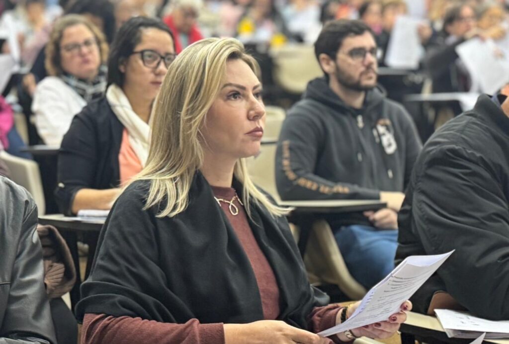 A imagem mostra uma sala de aula ou auditório lotado de pessoas sentadas em fileiras de cadeiras. O foco principal é uma mulher com cabelos loiros e compridos, vestindo uma blusa marrom-avermelhada e um casaco preto. Ela está segurando alguns papéis e parece estar atenta a algo. Ao seu redor, há outras pessoas, incluindo uma mulher com óculos e cabelos escuros, um homem com um casaco de moletom preto e outras pessoas com rostos parcialmente visíveis. O ambiente parece ser um evento formal ou acadêmico. A iluminação é suave e a qualidade da imagem é boa, permitindo uma visão clara dos detalhes. A composição da imagem é naturalista, com as pessoas sentadas em posições variadas e sem uma pose artificial. A imagem transmite uma sensação de atenção e envolvimento, sugerindo que as pessoas estão participando de uma palestra, apresentação ou outro evento semelhante.