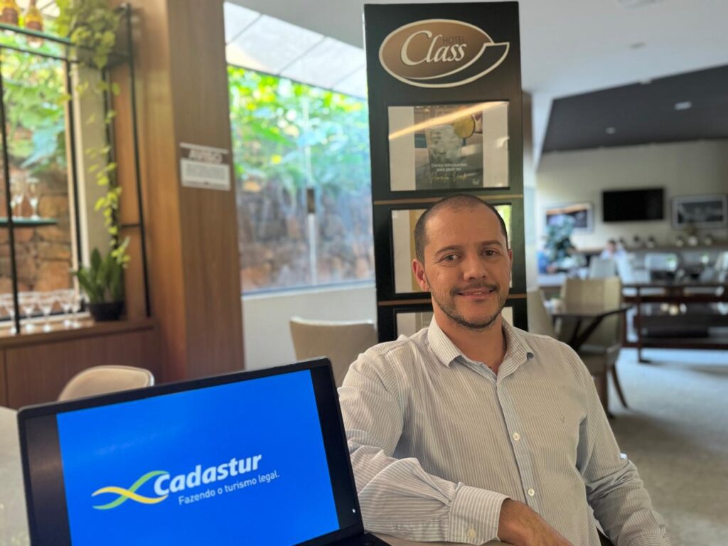 A imagem mostra um homem de meia-idade sentado em uma cadeira em um hotel ou lobby de hotel. Ele está vestindo uma camisa social azul clara e parece estar relaxado e confortável. Ele está olhando diretamente para a câmera com um sorriso leve no rosto.Ao fundo, há um ambiente de hotel, com uma recepção, uma televisão de tela plana na parede, outras pessoas sentadas em sofás e cadeiras, e algumas plantas. Há uma placa que diz "Hotel Class" atrás dele. Na frente dele, há um laptop aberto mostrando o logotipo da Cadastur, uma empresa que trabalha com o turismo.A imagem é bem iluminada e nítida, com cores vibrantes e um foco claro no homem sentado. O ambiente parece luxuoso e acolhedor. A imagem provavelmente foi tirada para um artigo de jornal ou blog, ou para fins de marketing, devido à presença do logotipo da Cadastur.Em resumo, a imagem transmite uma sensação de profissionalismo, conforto e elegância, representando possivelmente uma entrevista ou um evento de negócios em um ambiente de hotel.