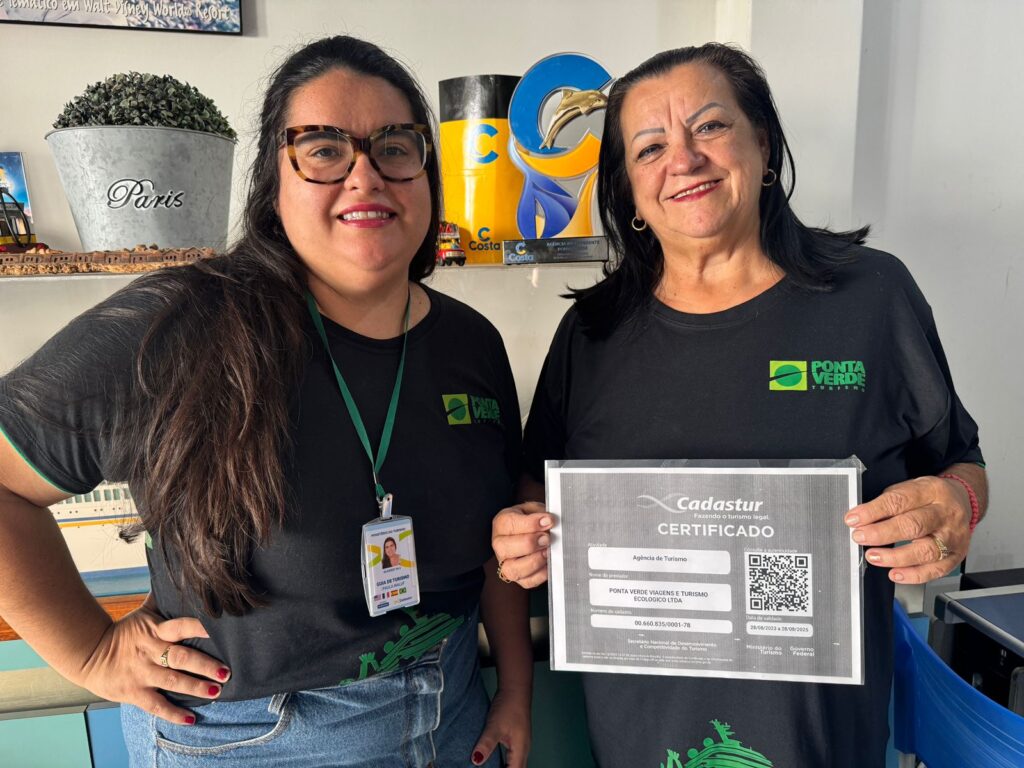 A imagem mostra duas mulheres sorrindo, segurando um certificado do Cadastur (Cadastro de Prestadores de Serviços Turísticos). Elas estão vestindo camisas pretas com o logotipo da "Ponta Verde Turismo". A mulher à esquerda tem um crachá de guia de turismo pendurado no pescoço.  Ao fundo, há uma prateleira com objetos decorativos, incluindo um balde com a palavra "Paris" escrita nele.  Há também uma placa de premiação da "Costa". O certificado mostra o nome da empresa "Ponta Verde Viagens e Turismo Ecológico Ltda" e seu número de cadastro. A data de validade do certificado também é visível. A imagem sugere que a empresa recebeu a certificação do Cadastur.