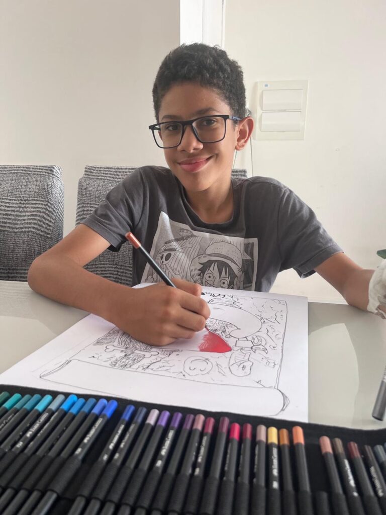 Menino negro de óculos, com camiseta cinza estampada, sorri enquanto desenha com lápis de cor sobre uma folha com esboço de ilustração. À frente, há um estojo com muitos lápis coloridos organizados. Ele está sentado em uma mesa branca, em um ambiente interno iluminado.