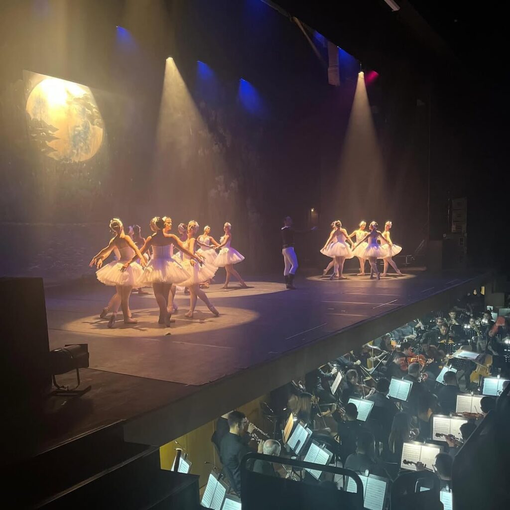 Em cena iluminada por luzes coloridas, várias bailarinas com tutus brancos dançam em formação circular. Ao fundo, um cenário com uma pintura de lua cheia. Abaixo do palco, vê-se a orquestra ao vivo, com músicos tocando instrumentos de cordas e sopros.