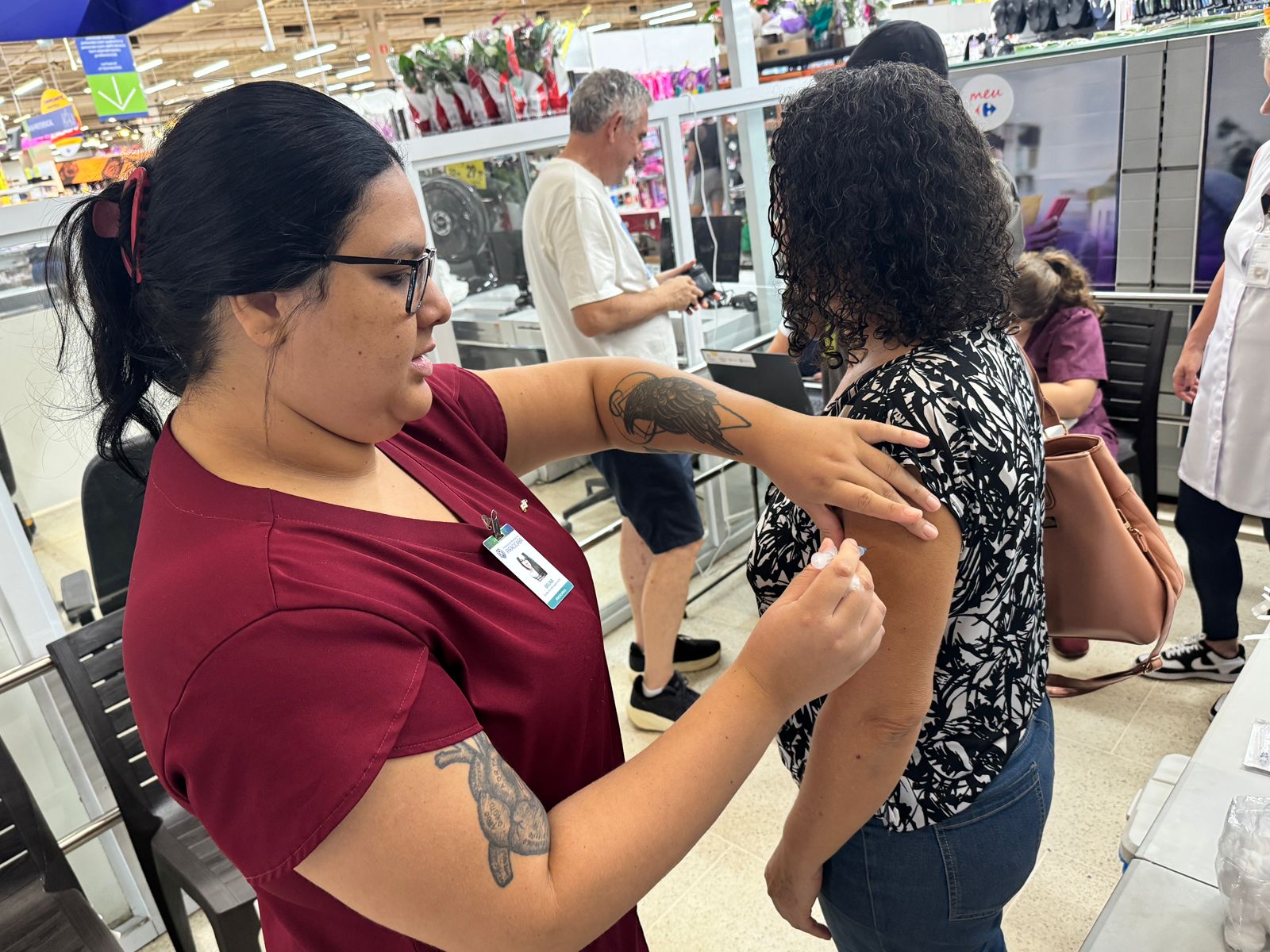 A imagem mostra uma profissional de saúde aplicando uma injeção no braço de uma mulher em um ambiente que parece ser uma farmácia ou supermercado. A profissional de saúde veste uma camiseta vinho escura com um crachá de identificação visível e tem tatuagens nos braços. A mulher que está recebendo a injeção veste uma blusa preta e branca com um padrão floral e jeans. Ela carrega uma bolsa marrom. Outras pessoas podem ser vistas ao fundo, algumas aparentemente esperando para serem atendidas. O ambiente parece organizado, mas movimentado. A cena sugere uma campanha de vacinação ou aplicação de medicamentos em um local público de fácil acesso. A imagem é nítida e bem iluminada, permitindo uma visão clara da ação principal.