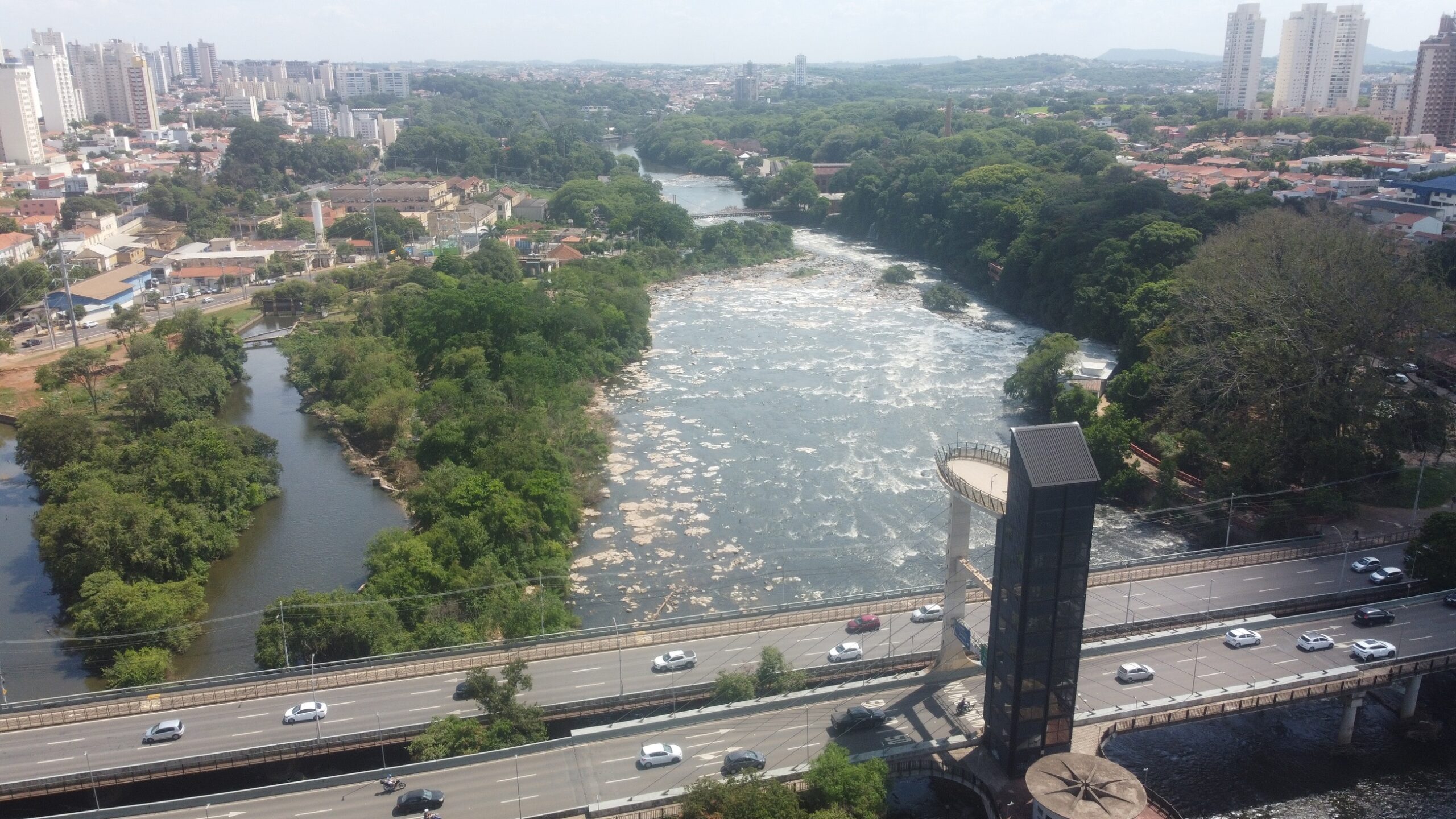 A imagem mostra uma vista aérea de uma parte de uma cidade, provavelmente no Brasil, com um rio correndo através dela. Vamos analisar a imagem em detalhes: *Elementos Geográficos:* * *Rio:* Um rio com corredeiras visíveis domina a parte central da imagem. A água parece turva, indicando possivelmente um rio de curso rápido ou com sedimentos. A presença de rochas expostas na superfície da água reforça essa ideia. Há uma bifurcação menor do rio, criando uma pequena ilha ou península coberta de vegetação. * *Vegetação:* A vegetação é abundante, principalmente ao longo das margens do rio e em áreas não urbanizadas. Apresenta-se como uma mistura de árvores de diferentes tamanhos, criando uma paisagem verdejante e densa. * *Urbanização:* A cidade se estende ao fundo e nas laterais da imagem. É possível observar edifícios de diferentes alturas, desde casas baixas até prédios altos, típicos de uma área urbana. A densidade da construção varia, com áreas mais densamente povoadas próximas ao rio e outras com construções mais espaçadas. *Elementos Construídos:* * *Ponte:* Uma ponte moderna de múltiplas pistas de rodagem atravessa o rio, sendo um elemento central da imagem. Ela possui um design que parece funcional e integrado à paisagem. Há uma estrutura vertical proeminente, possivelmente uma torre de observação ou parte da estrutura da ponte, com um mirante circular na parte superior. * *Estradas:* Estradas bem definidas e movimentadas são visíveis, principalmente as que compõem a ponte e as que a conectam à cidade. O trânsito de veículos é perceptível, indicando uma área com fluxo de carros significativo. * *Edifícios:* Como mencionado, os edifícios variam em tamanho e estilo arquitetônico, refletindo a variedade de usos do solo na cidade. Há prédios de múltiplos andares, indicando áreas residenciais ou comerciais. *Interpretação Geral:* A imagem sugere uma cidade em desenvolvimento, com uma boa integração entre a área urbana e o ambiente natural. A presença do rio e da vegetação contribui para a beleza cênica, enquanto a ponte e as estradas mostram a infraestrutura que conecta a cidade. A estrutura vertical na ponte pode ser um ponto turístico,