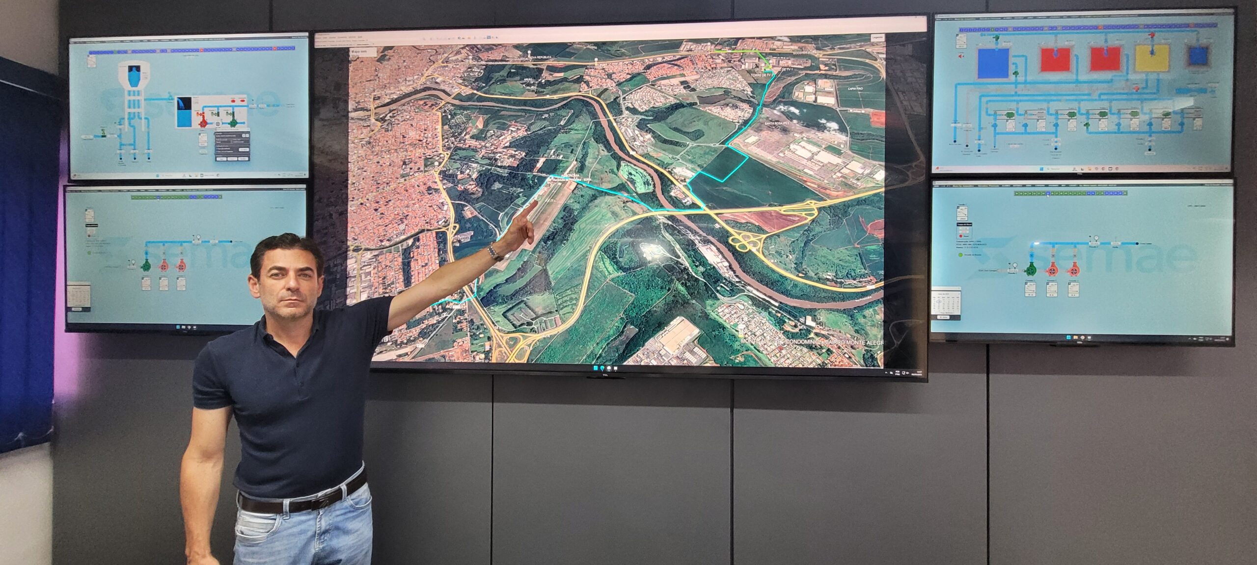 A imagem mostra um homem em pé na frente de uma parede com várias telas. A tela central, a maior, exibe um mapa aéreo de uma área urbana, com um trajeto destacado em azul claro. As telas menores, posicionadas em pares acima e abaixo da tela central, mostram diagramas que parecem representar fluxogramas de um processo, possivelmente industrial ou de infraestrutura, mostrando equipamentos e o fluxo entre eles. Os diagramas usam códigos de cores e símbolos para indicar diferentes estágios ou componentes do processo. A imagem sugere que o homem está apresentando ou explicando algo relacionado ao sistema representado nos diagramas e no mapa, possivelmente um sistema de monitoramento ou controle. A imagem não fornece informações suficientes para determinar o contexto específico, mas indica um ambiente de controle e monitoramento de um processo complexo.