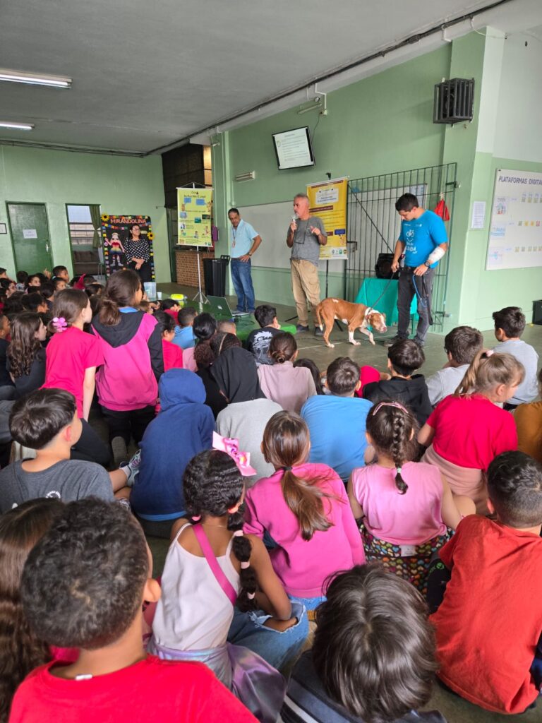 A imagem mostra uma apresentação em uma escola, provavelmente para crianças do ensino fundamental. Há dois homens adultos falando para uma grande assembleia de crianças sentadas no chão. Um deles está usando um microfone. Há também um terceiro homem adulto segurando uma guia de um cachorro marrom que está sentado calmamente perto deles. No fundo, há um quadro branco com uma apresentação de slides e cartazes informativos, sugerindo um tema educativo. Há também uma garota vestida de forma chamativa em um canto, possivelmente parte de outra apresentação ou atividade paralela. A atmosfera parece ser calma e atenciosa, com as crianças aparentemente engajadas na apresentação. O ambiente é uma sala de aula ou um espaço semelhante dentro de uma escola, com paredes verdes e um teto simples. A imagem sugere uma sessão de educação ou conscientização sobre um tema que envolve animais, possivelmente cães, dado a presença do cão na apresentação.