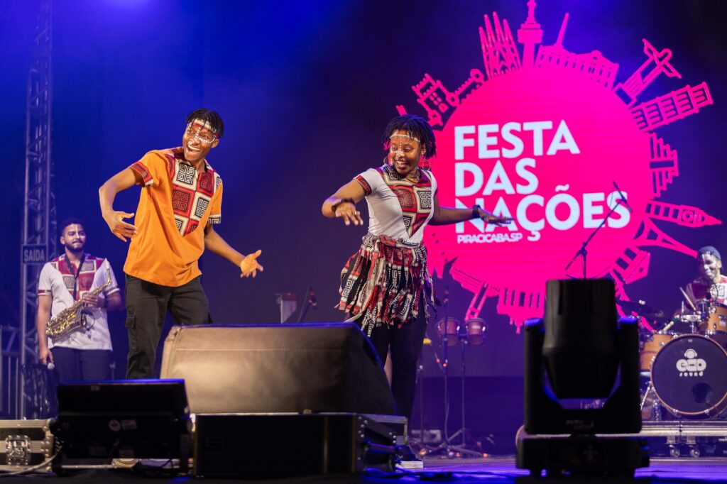 A imagem captura um momento vibrante de uma apresentação ao vivo no palco da Festa das Nações em Piracicaba, SP. O fundo do palco exibe um grande círculo rosa com o nome do evento. No primeiro plano, dois dançarinos em trajes tradicionais estampados com padrões geométricos realizam movimentos expressivos—um vestindo uma camisa laranja com calça preta e outro com uma blusa branca e saia estampada. Ao fundo, músicos tocam instrumentos como saxofone e tambores, complementando a energia festiva da cena. A iluminação e o equipamento de som profissional destacam o espetáculo, criando um ambiente cultural animado. Parece ser um evento repleto de celebração, diversidade e homenagens às tradições de diferentes nações.