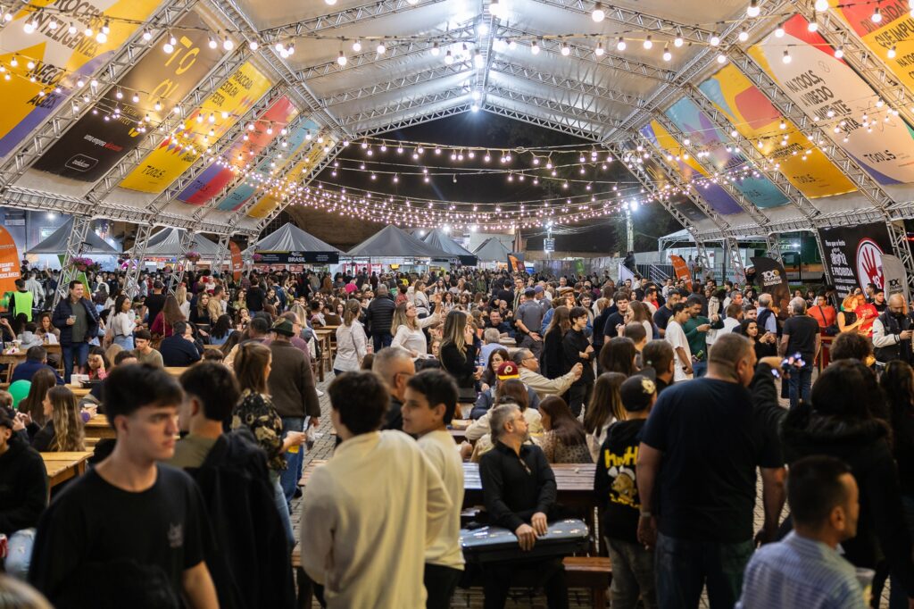 A imagem mostra um grande público reunido sob uma tenda iluminada por fileiras de luzes na Festa das Nações de Piracicaba. Pessoas de todas as idades circulam, conversam e aproveitam pratos típicos nos estandes ao fundo. O clima é animado e familiar. O espaço está decorado com painéis coloridos e mensagens culturais.