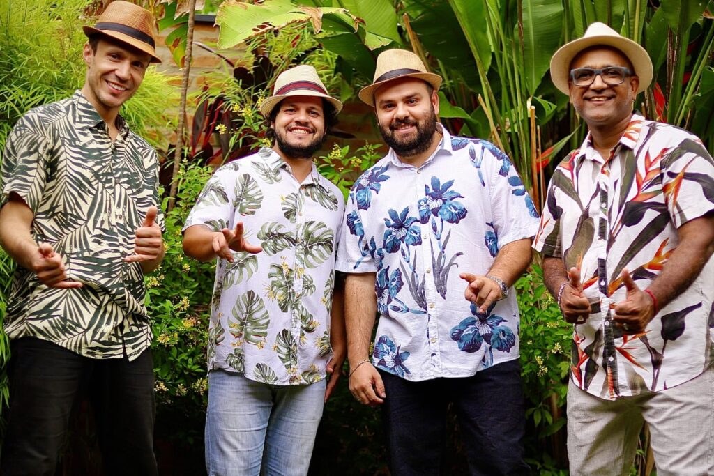 Quatro homens estão posicionados lado a lado, sorrindo, em um ambiente ao ar livre cercado por vegetação tropical com folhas grandes e verdes. Todos vestem camisas estampadas com folhagens ou flores e usam chapéus de palha estilo Panamá. O grupo transmite um clima alegre e descontraído, típico de apresentações musicais brasileiras. Ao fundo, plantas exuberantes complementam o cenário natural. A imagem é promocional do grupo musical Brasil Mestiço.