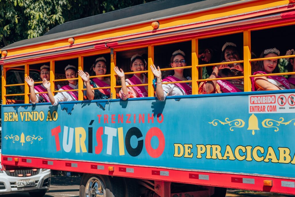 A imagem mostra um animado trenzinho turístico em Piracicaba, repleto de passageiros sorridentes que acenam das janelas. O trem é vibrante e decorado com um cartaz de boas-vindas que diz: "Bem Vindo Ao Trenzinho Turístico de Piracicaba." Dentro do trem, algumas pessoas usam faixas e tiaras, indicando que podem estar participando de um evento especial ou celebração.