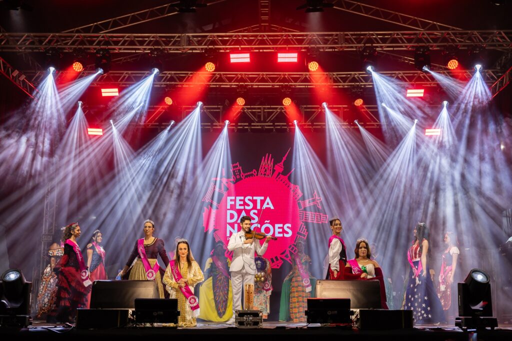 A imagem mostra um palco iluminado com luzes vibrantes, onde um grupo de pessoas veste trajes tradicionais e coloridos. No centro, há um grande painel circular com o texto "FESTA DAS NAÇÕES", indicando que o evento é uma celebração cultural com representações de diferentes países. Algumas das pessoas no palco estão usando faixas, sugerindo que podem ser participantes de um concurso ou desfile temático. O ambiente parece festivo e cheio de diversidade cultural.
