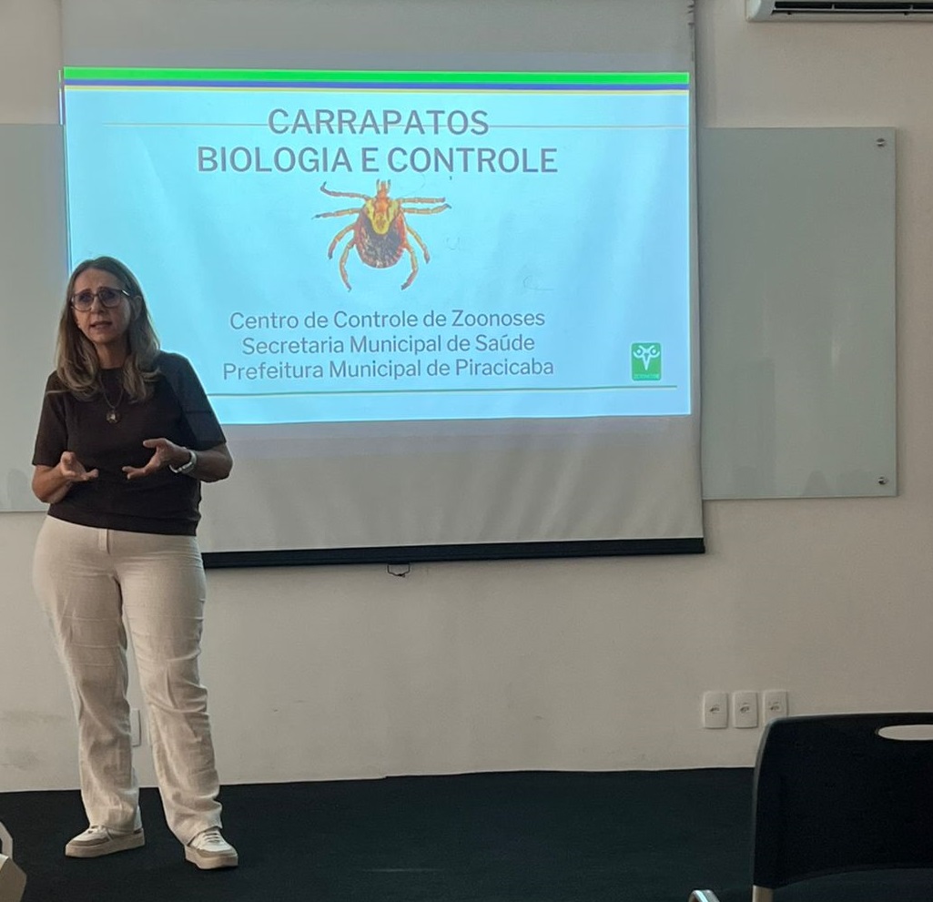 A imagem mostra uma apresentação sobre carrapatos, mais especificamente sobre sua biologia e controle. A apresentação é dada por uma palestrante, uma mulher de meia idade, vestindo roupas sociais, em frente a uma tela de projeção. A tela mostra o título "CARRAPATOS BIOLOGIA E CONTROLE", acompanhado de uma imagem de um carrapato. Na parte inferior, há o rodapé indicando a autoria da apresentação: "Centro de Controle de Zoonoses, Secretaria Municipal de Saúde, Prefeitura Municipal de Piracicaba". A presença do logotipo da prefeitura reforça a origem oficial da apresentação. A imagem sugere que a apresentação está ocorrendo em um ambiente formal, possivelmente uma sala de conferências ou de aula, dado o cenário e a natureza da apresentação. A palestrante parece envolvida e engajada com o conteúdo, sugerindo uma apresentação informativa e possivelmente educativa sobre os carrapatos, seus ciclos de vida, e métodos de controle. A apresentação provavelmente se destina a um público interessado em saúde pública ou controle de pragas.
