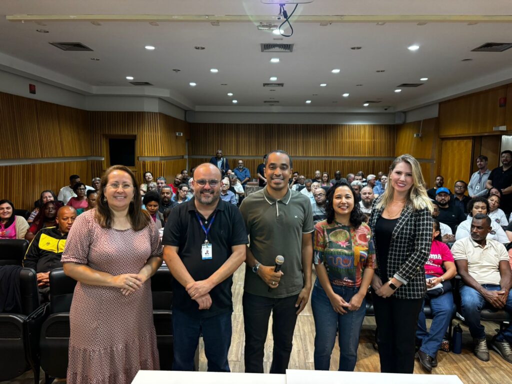 A imagem mostra um grupo de pessoas em uma sala de conferência ou reunião. Há cinco indivíduos em pé na frente da sala, com uma pessoa segurando um microfone. O ambiente está preenchido com participantes sentados, sugerindo um evento formal, como uma apresentação, seminário ou discussão. A sala tem painéis de madeira nas paredes e um projetor montado no teto.