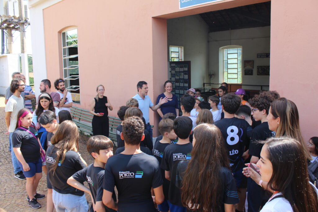A imagem mostra um grande grupo de pessoas reunidas na entrada de um prédio de tijolo cor de rosa. Parece ser uma excursão escolar ou um evento comunitário. *As pessoas:* * *À frente:* Duas pessoas, um homem e uma mulher, estão falando para o grupo. Eles parecem ser professores, guias ou palestrantes. * *No meio:* Um grande grupo de crianças e adolescentes está reunido, muitos vestindo camisetas pretas com o logotipo "Primeiro Porto". Pode ser uma escola ou organização. * *Na parte de trás:* Vários adultos estão observando, alguns parecendo pais ou responsáveis. *O ambiente:* * *O prédio:* É um prédio de tijolo cor de rosa, antigo, com janelas de madeira e uma entrada coberta. Há uma placa acima da entrada, mas o texto não é legível. Há também algumas imagens na parede interna. * *O exterior:* O exterior é simples, com arbustos e uma cerca visíveis ao fundo. *A atmosfera:* A atmosfera é amigável e informativa. Parece ser uma apresentação ou discussão, com o grupo prestando atenção aos palestrantes. *Possíveis interpretações:* A imagem poderia representar uma aula ao ar livre, uma visita guiada a um local histórico, ou uma apresentação de um projeto escolar. A presença do logotipo "Primeiro Porto" sugere uma conexão com uma escola ou organização com esse nome. Em resumo, a imagem captura um momento de aprendizagem e interação social em um ambiente histórico. A uniformidade das camisetas sugere uma identidade de grupo, provavelmente uma escola ou organização. A conversa entre as pessoas na frente sugere uma apresentação.