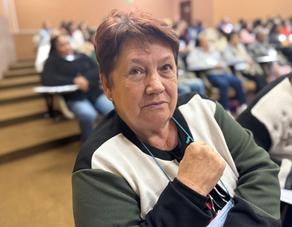 A imagem mostra uma mulher de meia-idade, com cabelo curto castanho-avermelhado e pele enrugada, sentada numa cadeira numa sala de conferências. Ela está vestida com um casaco de três cores, com mangas pretas, uma parte branca no meio e uma parte verde-oliva na parte de fora. Ela segura o punho direito fechado, e o seu olhar é direto para a câmara, com uma expressão séria e ligeiramente séria.O fundo está desfocado, mas mostra uma sala de conferências cheia de outras pessoas sentadas em cadeiras, sugerindo uma reunião ou apresentação. As cadeiras são de madeira e organizadas em filas. A iluminação é suave e difusa, sem sombras duras. A imagem é nítida e bem iluminada, com foco na mulher em primeiro plano.