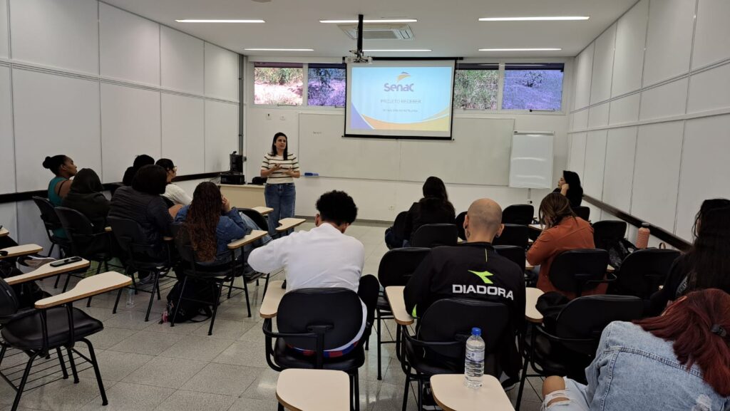 A imagem mostra uma sala de aula com uma palestrante apresentando uma apresentação de PowerPoint para uma turma de alunos.*Detalhes:** *A palestrante:* Uma mulher está de pé na frente da sala, apresentando uma apresentação no PowerPoint com o logotipo do Senac e o título "Projeto Receber". Ela parece confiante e bem vestida, usando uma blusa de manga comprida e calças jeans.* *A apresentação:* A apresentação no PowerPoint exibe o logotipo do Senac e o título "Projeto Receber". Há também algum texto adicional na apresentação, mas é ilegível na imagem.* *Os alunos:* A sala de aula está cheia de alunos sentados em carteiras individuais. Eles parecem estar prestando atenção na palestrante. Os alunos são uma mistura de homens e mulheres, com diversas idades e etnias. Alguns alunos estão sentados sozinhos, enquanto outros estão em grupos.* *O ambiente:* A sala de aula é moderna e bem iluminada. As paredes são brancas e há uma janela grande que mostra a vegetação externa. A sala é organizada e limpa. Há um quadro branco e um projetor na sala.* *O clima:* O clima parece ser formal e profissional. A palestrante parece estar conduzindo uma apresentação profissional, e os alunos parecem estar envolvidos no assunto.Em resumo, a imagem mostra uma apresentação profissional em uma sala de aula do Senac. A palestrante está apresentando um projeto chamado "Projeto Receber" para uma turma atenta de alunos. O ambiente é limpo, moderno e profissional.