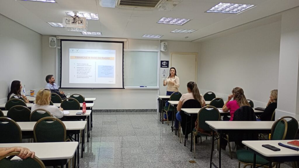 A imagem mostra uma sala de aula com várias pessoas sentadas em mesas e cadeiras, ouvindo uma palestrante. A palestrante está de pé na frente de uma tela de projeção, na qual é possível ver uma apresentação de slides sobre "Finanças e Fases da Razão".A sala de aula é bem iluminada e organizada, com mesas e cadeiras dispostas em fileiras. As paredes são claras e o chão é de um material liso e claro, provavelmente cerâmica ou piso laminado.  Há um quadro branco vazio na parede ao lado da tela de projeção. Um projetor está pendurado no teto.As pessoas na sala parecem estar atentas à apresentação. A palestrante parece confiante e engajada. A atmosfera parece ser profissional e calma. Há um sinal de Wi-Fi visível na parede.  A imagem sugere uma apresentação ou workshop sobre finanças.Em resumo, a imagem retrata uma cena de uma apresentação ou aula sobre finanças em um ambiente de sala de aula formal e profissional.