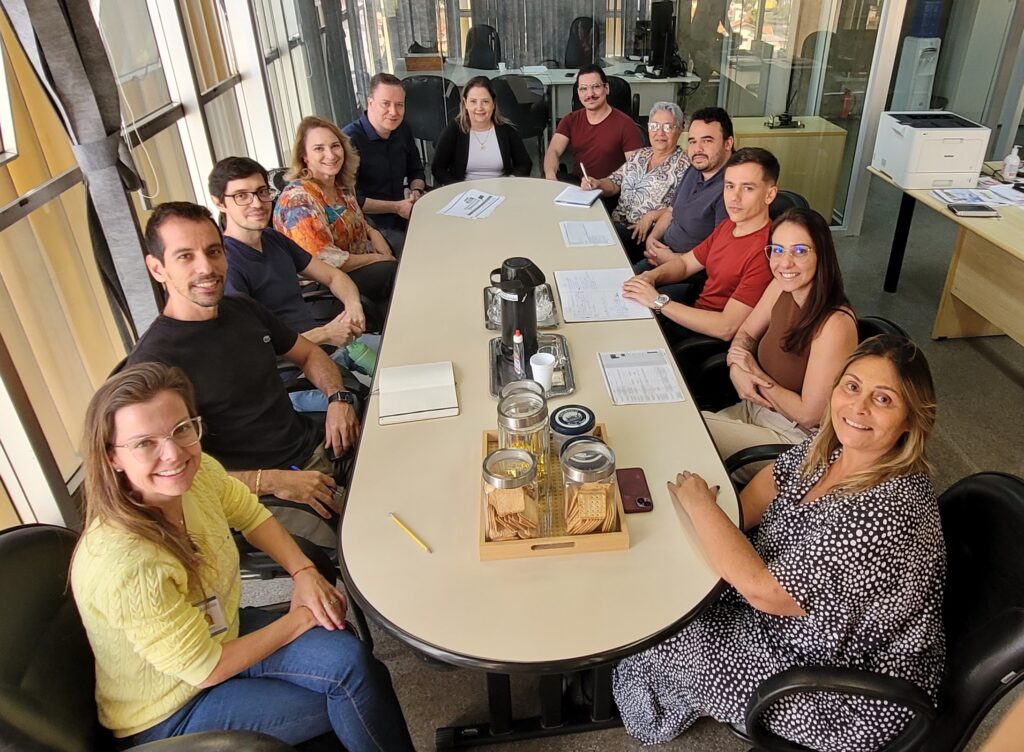 A foto mostra um grupo de 14 pessoas sentadas em torno de uma mesa oval em uma sala de reuniões. A sala parece ser um escritório, com janelas grandes que deixam entrar muita luz natural. A mesa está coberta com documentos, canetas, uma cafeteira e alguns biscoitos. As pessoas na foto parecem estar em uma reunião, algumas estão olhando para a câmera, outras estão envolvidas em conversas. Há uma mistura de homens e mulheres, com idades variadas. Eles parecem ser um grupo diverso, com diferentes origens étnicas e estilos de vestuário. O ambiente geral parece ser profissional e amigável. A imagem sugere que a reunião é importante, dado o número de pessoas presentes e a quantidade de papéis na mesa. A luz natural e a disposição das pessoas contribuem para uma atmosfera acolhedora e produtiva. A foto provavelmente foi tirada para documentar a reunião ou para capturar um momento entre colegas de trabalho.