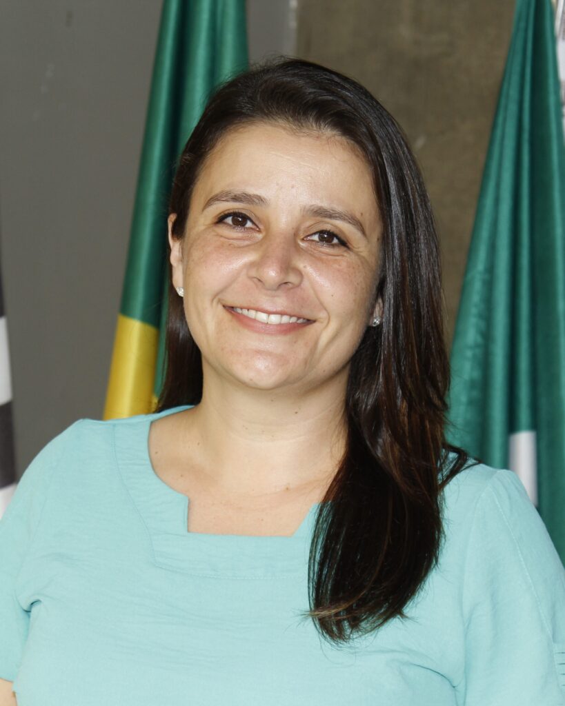 A imagem mostra um retrato de uma mulher de meia-idade com cabelo castanho escuro e pele clara. Ela está vestindo uma blusa verde-água de gola em V. Ela está sorrindo levemente para a câmera. No fundo, há duas bandeiras parcialmente visíveis, sugerindo um ambiente formal ou governamental. A mulher parece confiante e calma. A iluminação é suave e difusa, sem sombras duras no rosto. A qualidade da imagem é boa, com detalhes nítidos e cores precisas. A composição é simples e direta, com o foco principal na mulher. A imagem transmite uma sensação de profissionalismo e serenidade.