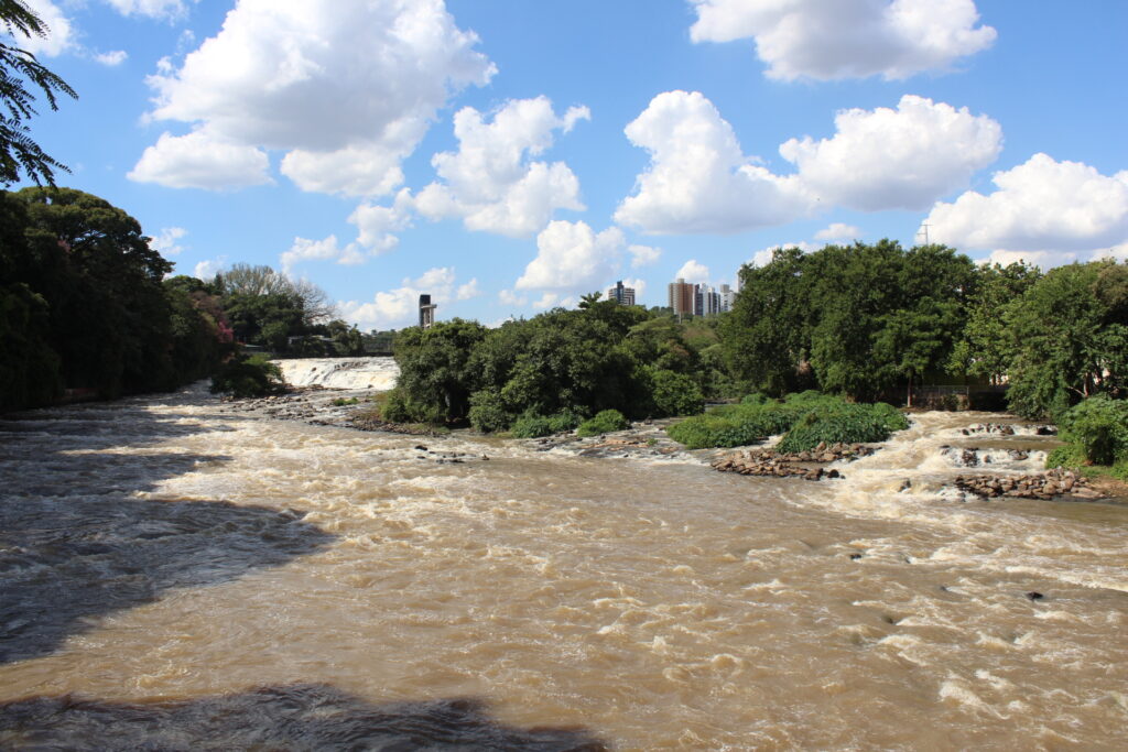 A imagem mostra um rio caudaloso com água turva, possivelmente devido a sedimentos ou alta vazão. A água está agitada, com pequenas corredeiras e quedas d'água visíveis em alguns pontos. A vegetação ao longo das margens é densa, composta principalmente por árvores de folhagem verde escura. Há um contraste entre a vegetação exuberante e a água barrenta. Ao fundo, é possível observar prédios de vários andares, indicando a proximidade de uma área urbana. O céu está parcialmente nublado, com nuvens brancas e volumosas contra um céu azul claro. A cena sugere um ambiente natural próximo a uma área urbanizada, possivelmente um rio que atravessa ou margeia uma cidade. A luz do dia indica que a foto foi tirada durante o dia, provavelmente em condições de tempo bom, mas não ensolarado. A perspectiva da foto é de um ponto elevado, possivelmente uma ponte ou um mirante.