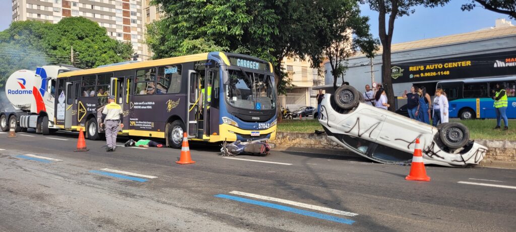 A imagem mostra a cena de um acidente de trânsito. Um carro branco está de capota no meio da rua, cercado por bombeiros e outros socorristas. Há um ônibus, aparentemente envolvido no acidente, parado próximo ao carro capotado. Uma ambulância do SAMU (Serviço de Atendimento Móvel de Urgência) também está presente no local. Diversas pessoas, incluindo bombeiros vestidos com uniformes, estão envolvidas no atendimento às vítimas e na remoção do veículo. A rua parece ser em uma área urbana, com lojas e prédios ao fundo. A placa de uma das lojas indica "ARICO" e outra diz "ALTO GIRO". Um detalhe interessante é a presença de um drone no céu, possivelmente registrando a ocorrência. O ônibus apresenta uma pintura com notas musicais e a inscrição "RITMO DO TRÂNSITO". A cena sugere um acidente grave que requer a intervenção de múltiplos serviços de emergência.