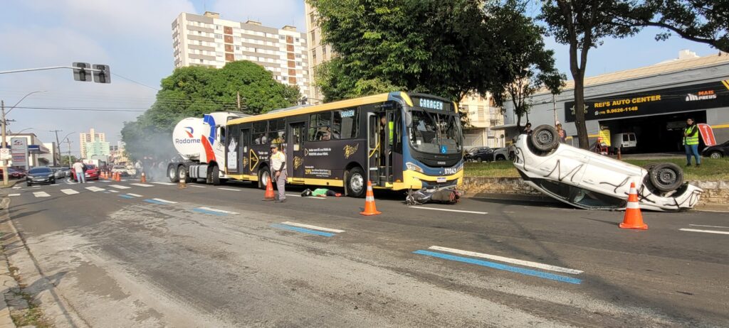 A imagem mostra a cena de um acidente de trânsito. Um carro branco está de capota no meio da rua, cercado por bombeiros e outros socorristas. Há um ônibus, aparentemente envolvido no acidente, parado próximo ao carro capotado. Uma ambulância do SAMU (Serviço de Atendimento Móvel de Urgência) também está presente no local. Diversas pessoas, incluindo bombeiros vestidos com uniformes, estão envolvidas no atendimento às vítimas e na remoção do veículo. A rua parece ser em uma área urbana, com lojas e prédios ao fundo. A placa de uma das lojas indica "ARICO" e outra diz "ALTO GIRO". Um detalhe interessante é a presença de um drone no céu, possivelmente registrando a ocorrência. O ônibus apresenta uma pintura com notas musicais e a inscrição "RITMO DO TRÂNSITO". A cena sugere um acidente grave que requer a intervenção de múltiplos serviços de emergência.