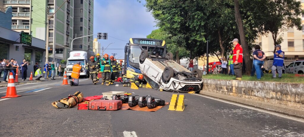 A imagem mostra a cena de um acidente de trânsito. Um carro branco está de capota no meio da rua, cercado por bombeiros e outros socorristas. Há um ônibus, aparentemente envolvido no acidente, parado próximo ao carro capotado. Uma ambulância do SAMU (Serviço de Atendimento Móvel de Urgência) também está presente no local. Diversas pessoas, incluindo bombeiros vestidos com uniformes, estão envolvidas no atendimento às vítimas e na remoção do veículo. A rua parece ser em uma área urbana, com lojas e prédios ao fundo. A placa de uma das lojas indica "ARICO" e outra diz "ALTO GIRO". Um detalhe interessante é a presença de um drone no céu, possivelmente registrando a ocorrência. O ônibus apresenta uma pintura com notas musicais e a inscrição "RITMO DO TRÂNSITO". A cena sugere um acidente grave que requer a intervenção de múltiplos serviços de emergência.