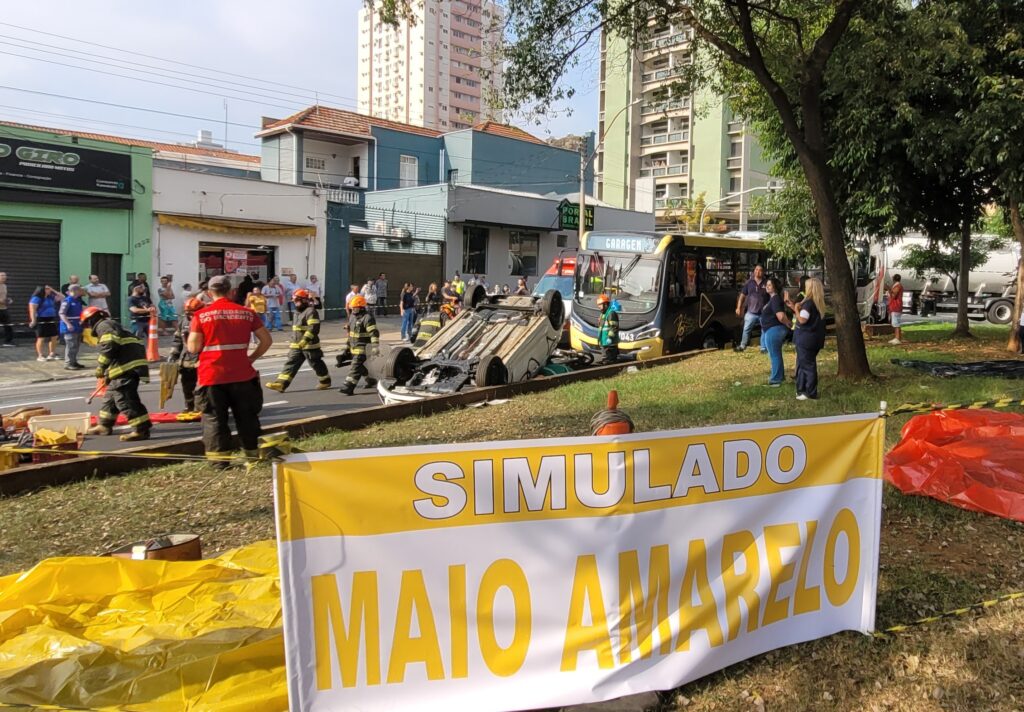 A imagem mostra a cena de um acidente de trânsito. Um carro branco está de capota no meio da rua, cercado por bombeiros e outros socorristas. Há um ônibus, aparentemente envolvido no acidente, parado próximo ao carro capotado. Uma ambulância do SAMU (Serviço de Atendimento Móvel de Urgência) também está presente no local. Diversas pessoas, incluindo bombeiros vestidos com uniformes, estão envolvidas no atendimento às vítimas e na remoção do veículo. A rua parece ser em uma área urbana, com lojas e prédios ao fundo. A placa de uma das lojas indica "ARICO" e outra diz "ALTO GIRO". Um detalhe interessante é a presença de um drone no céu, possivelmente registrando a ocorrência. O ônibus apresenta uma pintura com notas musicais e a inscrição "RITMO DO TRÂNSITO". A cena sugere um acidente grave que requer a intervenção de múltiplos serviços de emergência.