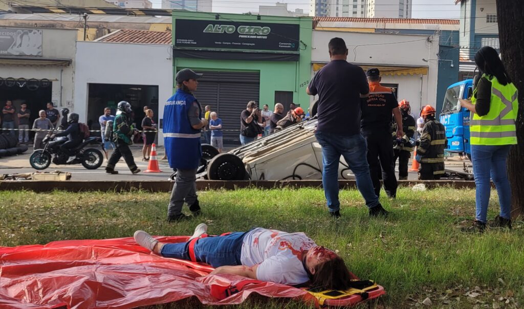 A imagem mostra a cena de um acidente de trânsito. Um carro branco está de capota no meio da rua, cercado por bombeiros e outros socorristas. Há um ônibus, aparentemente envolvido no acidente, parado próximo ao carro capotado. Uma ambulância do SAMU (Serviço de Atendimento Móvel de Urgência) também está presente no local. Diversas pessoas, incluindo bombeiros vestidos com uniformes, estão envolvidas no atendimento às vítimas e na remoção do veículo. A rua parece ser em uma área urbana, com lojas e prédios ao fundo. A placa de uma das lojas indica "ARICO" e outra diz "ALTO GIRO". Um detalhe interessante é a presença de um drone no céu, possivelmente registrando a ocorrência. O ônibus apresenta uma pintura com notas musicais e a inscrição "RITMO DO TRÂNSITO". A cena sugere um acidente grave que requer a intervenção de múltiplos serviços de emergência.