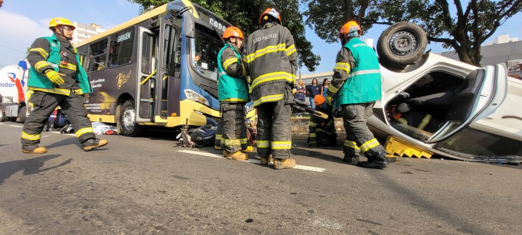 A imagem mostra a cena de um acidente de trânsito. Um carro branco está de capota no meio da rua, cercado por bombeiros e outros socorristas. Há um ônibus, aparentemente envolvido no acidente, parado próximo ao carro capotado. Uma ambulância do SAMU (Serviço de Atendimento Móvel de Urgência) também está presente no local. Diversas pessoas, incluindo bombeiros vestidos com uniformes, estão envolvidas no atendimento às vítimas e na remoção do veículo. A rua parece ser em uma área urbana, com lojas e prédios ao fundo. A placa de uma das lojas indica "ARICO" e outra diz "ALTO GIRO". Um detalhe interessante é a presença de um drone no céu, possivelmente registrando a ocorrência. O ônibus apresenta uma pintura com notas musicais e a inscrição "RITMO DO TRÂNSITO". A cena sugere um acidente grave que requer a intervenção de múltiplos serviços de emergência.