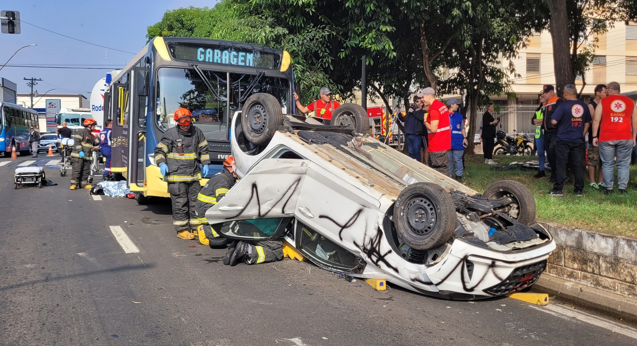 A imagem mostra a cena de um acidente de trânsito. Um carro branco está de capota no meio da rua, cercado por bombeiros e outros socorristas. Há um ônibus, aparentemente envolvido no acidente, parado próximo ao carro capotado. Uma ambulância do SAMU (Serviço de Atendimento Móvel de Urgência) também está presente no local. Diversas pessoas, incluindo bombeiros vestidos com uniformes, estão envolvidas no atendimento às vítimas e na remoção do veículo. A rua parece ser em uma área urbana, com lojas e prédios ao fundo. A placa de uma das lojas indica "ARICO" e outra diz "ALTO GIRO". Um detalhe interessante é a presença de um drone no céu, possivelmente registrando a ocorrência. O ônibus apresenta uma pintura com notas musicais e a inscrição "RITMO DO TRÂNSITO". A cena sugere um acidente grave que requer a intervenção de múltiplos serviços de emergência.