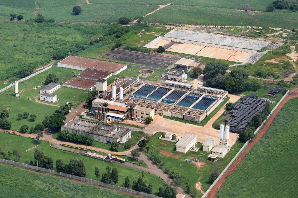 A imagem mostra uma vista aérea de uma instalação industrial, provavelmente uma usina de açúcar e álcool, rodeada por plantações de cana-de-açúcar. A imagem é detalhada o suficiente para permitir uma análise de vários aspectos da instalação: *Elementos principais da imagem:* * *Usina de açúcar e álcool (principal):* A parte central da imagem é dominada por uma grande estrutura que parece ser a usina principal. Ela inclui edifícios de processamento, tanques de armazenamento (provavelmente para o caldo de cana, álcool ou outros subprodutos), e equipamentos. A presença de silos e torres de armazenamento sugere atividades de processamento e armazenamento de grandes volumes de matéria-prima e produtos. * *Estação de tratamento de água (ETA):* Há uma seção claramente identificável como uma ETA, com vários tanques retangulares, que provavelmente são lagoas de tratamento ou decantadores. A disposição organizada dos tanques indica um processo de tratamento em etapas. * *Edifícios auxiliares:* Vários edifícios menores estão espalhados pela área, que provavelmente servem como escritórios administrativos, alojamentos, oficinas de manutenção ou outras funções de suporte. * *Armazenamento de materiais:* Uma área mostra uma pilha organizada de tubos ou vigas longas, provavelmente para uso na manutenção ou expansão da instalação. * *Cana-de-açúcar:* A instalação está cercada por extensos campos de cana-de-açúcar, indicando que a matéria-prima é produzida localmente. * *Infraestrutura:* A área possui estradas internas, muros, e outras infraestruturas necessárias para a operação da usina. *Inferências:* * *Escala:* A extensão da instalação sugere uma operação de grande porte, capaz de processar grandes quantidades de cana-de-açúcar. * *Integração:* A proximidade da ETA à usina principal indica uma integração eficiente do tratamento de água no processo industrial. A usina provavelmente usa grandes quantidades de água no processo de produção, necessitando de um sistema de tratamento de efluentes. * *Localização:* A presença de extensos campos de cana-de-açúcar sugere uma região com clima e solo apropriados para o cultivo dessa planta, provavelmente em uma zona rural.