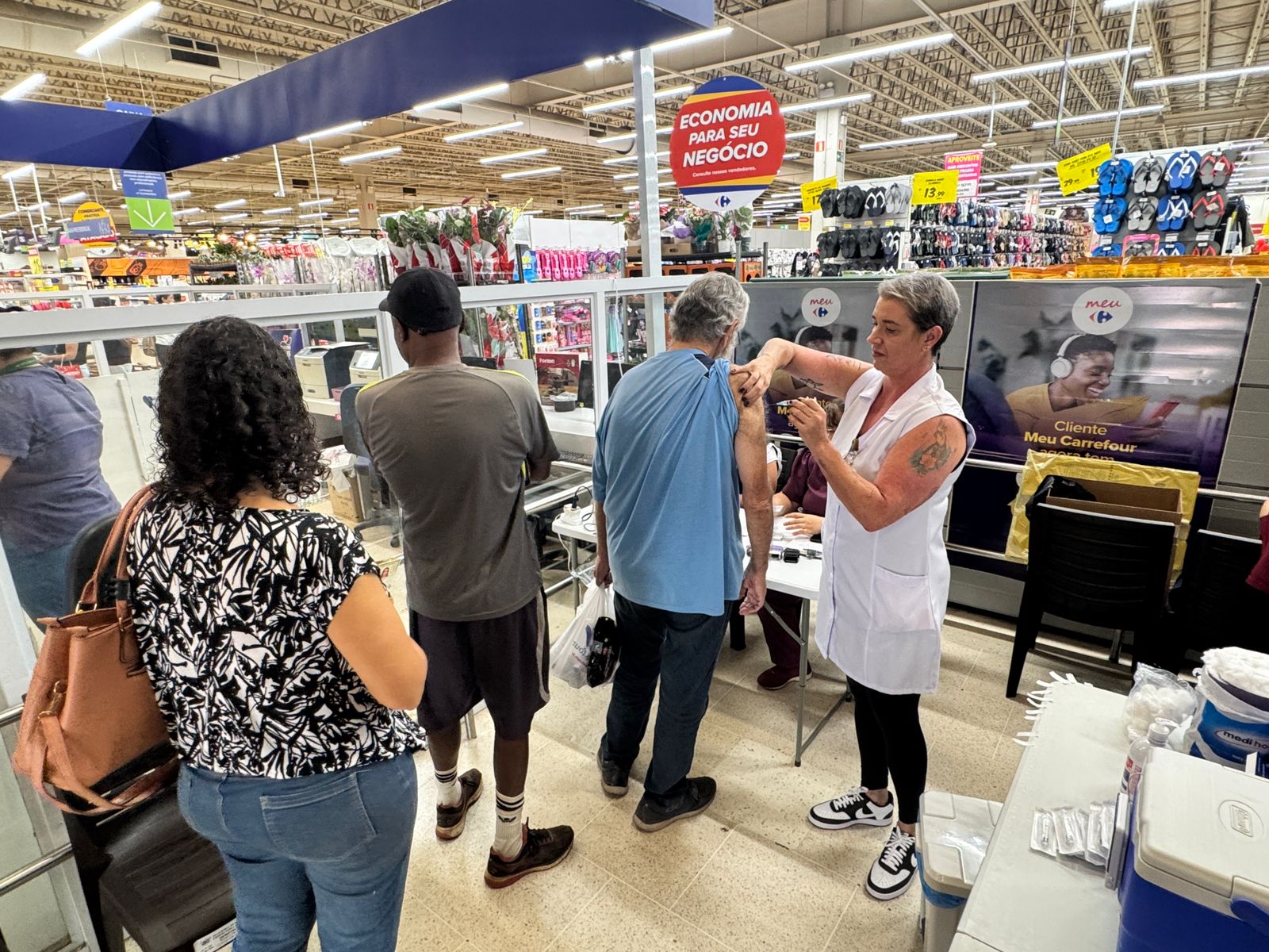 A imagem mostra uma campanha de vacinação acontecendo dentro de um supermercado Carrefour. Uma profissional de saúde, vestindo um jaleco branco, está aplicando uma injeção no braço de um homem. Há outras pessoas na fila esperando para serem vacinadas. A cena se passa em um corredor do supermercado, com prateleiras de produtos visíveis ao fundo. Há uma placa acima do local de vacinação que diz "Economia para seu negócio", indicando uma possível promoção ou parceria entre o Carrefour e a campanha de vacinação. Também há cartazes com a marca "Meu Carrefour", sugerindo uma iniciativa de bem-estar para os clientes. O ambiente parece organizado, com divisórias para separar os indivíduos na fila e materiais de primeiros socorros visíveis próximos à profissional de saúde. A imagem sugere um esforço para facilitar o acesso à vacinação, oferecendo-a em um local de conveniência para a população.