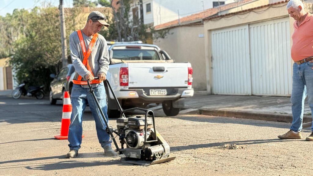 A imagem mostra dois indivíduos realizando um trabalho de reparo em uma estrada. Um deles está operando um compactador de placa, uma máquina usada para compactar solo, asfalto ou outros materiais, criando uma superfície nivelada. O outro está em pé observando o trabalho. Um cone de trânsito está colocado na via, indicando que a área está em manutenção. No fundo, há uma caminhonete branca e uma motocicleta estacionada ao lado da estrada.