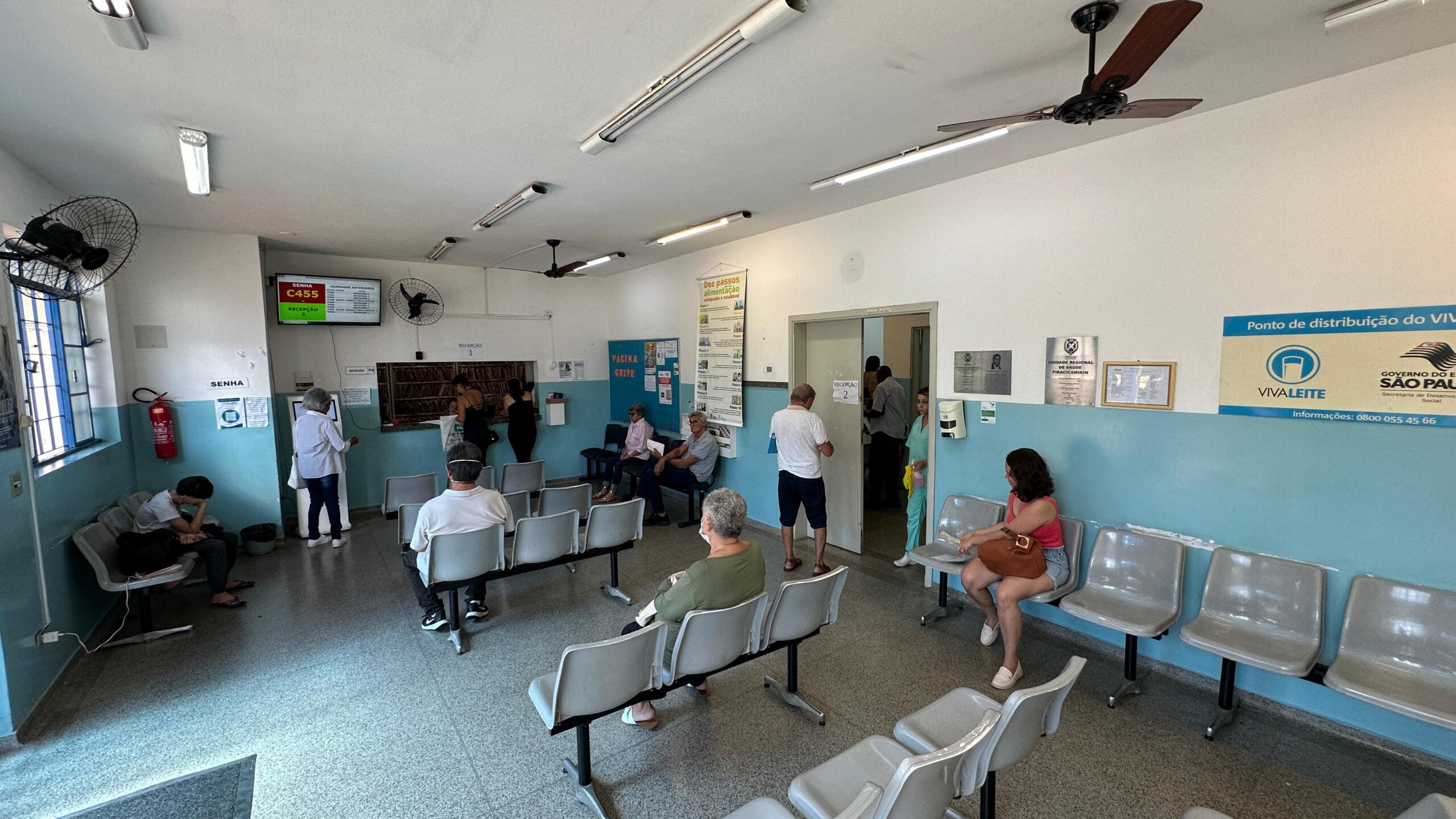 A imagem mostra a sala de espera de um posto de saúde ou clínica. Há várias pessoas sentadas em cadeiras de plástico cinzas, aguardando atendimento. Algumas pessoas estão usando máscaras. Há uma recepção com balcão e guichês, onde aparentemente se retiram senhas para atendimento. Uma tela exibe o número da senha atual (C455). Há também cartazes informativos sobre vacinação contra gripe e alimentação saudável. Na parede, há uma placa indicando um ponto de distribuição do programa VivaLeite, do governo do estado de São Paulo. O ambiente é simples, com ventiladores de teto e iluminação fluorescente. Nota-se a presença de um extintor de incêndio e dispensador de álcool em gel. Em resumo, a imagem retrata uma cena cotidiana de espera por atendimento médico em um estabelecimento público de saúde.