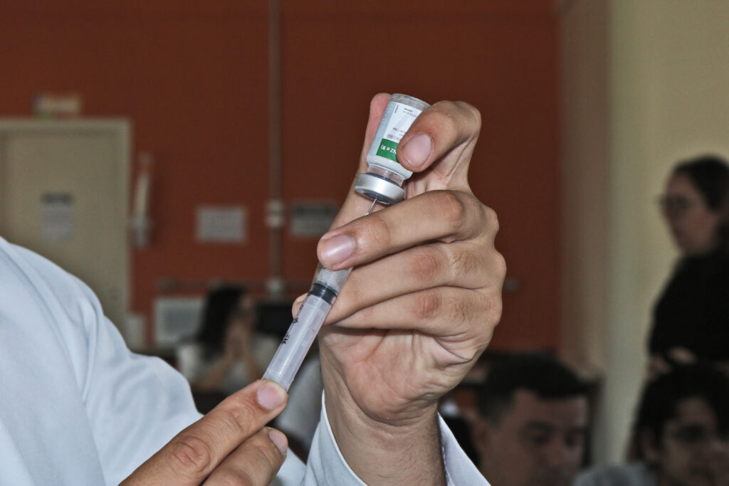 A imagem mostra um close de uma pessoa, aparentemente um profissional de saúde, preparando uma seringa para injeção. A pessoa está vestindo um jaleco branco e segura uma ampola pequena com uma mão, enquanto com a outra segura uma seringa, aparentemente a enchendo com o conteúdo da ampola. O fundo é desfocado, mas mostra uma sala com outras pessoas ao fundo, sugerindo um ambiente clínico ou médico, possivelmente um posto de saúde ou hospital. A imagem sugere um procedimento de vacinação ou outra forma de injeção médica. Não há detalhes suficientes para identificar o tipo de medicamento ou vacina. A qualidade da imagem é boa o suficiente para mostrar os detalhes do processo de preparação da injeção.