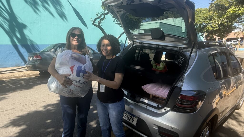 A imagem mostra duas mulheres em pé perto de um carro de passageiros. Uma mulher está segurando uma sacola plástica grande com itens não identificados. A outra mulher está ao lado dela, com os braços cruzados. O carro está aberto, mostrando seu porta-malas, que está parcialmente cheio de itens como travesseiros e brinquedos de pelúcia. As mulheres parecem estar envolvidas em alguma forma de atividade de caridade ou doação. O ambiente parece ser externo em um dia ensolarado. O fundo inclui uma parede azul e algumas árvores.